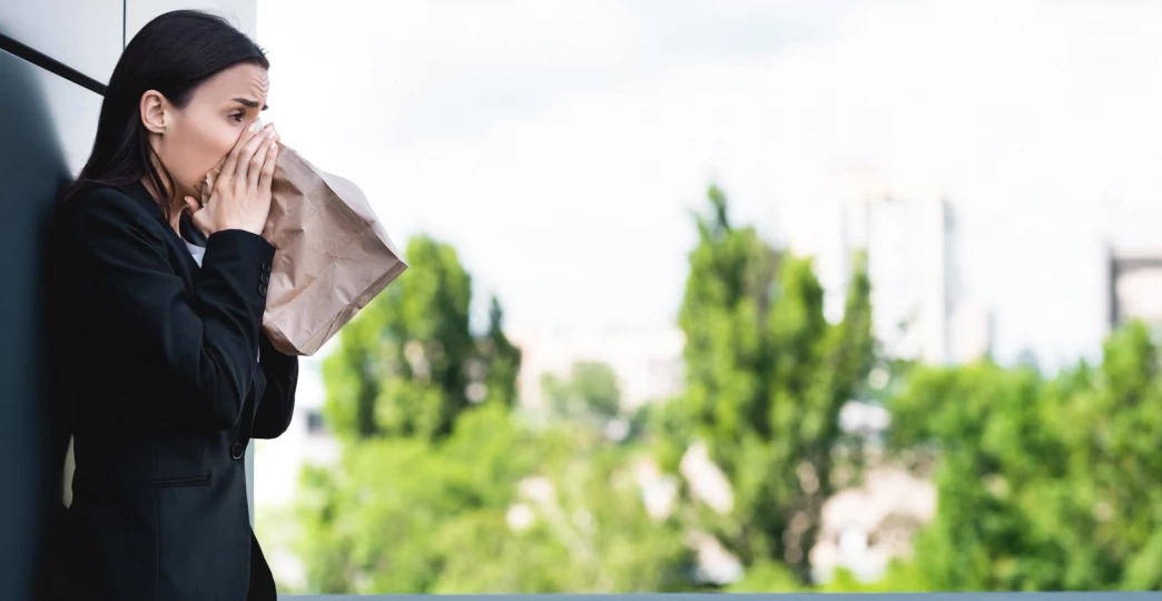 young businesswoman standing on rooftop and breathing into paper bag while suffering from panic