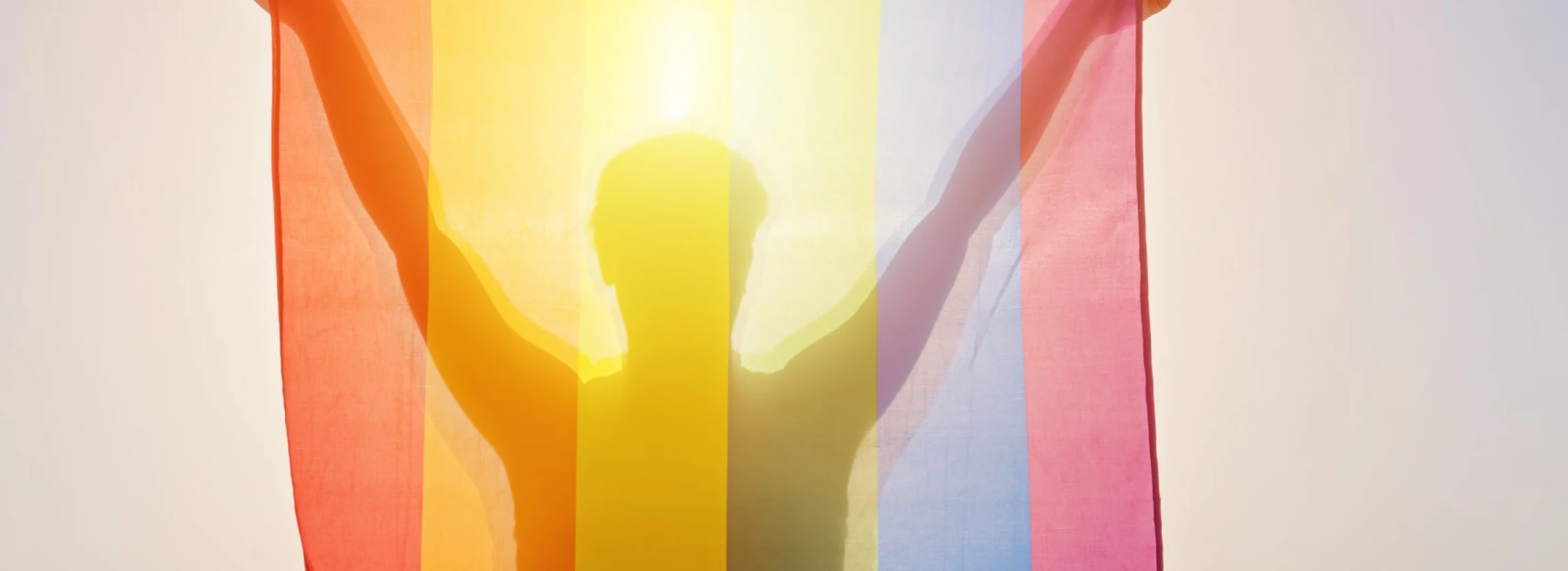 young woman holding raised hands waving LGBT rainbow flag against sky